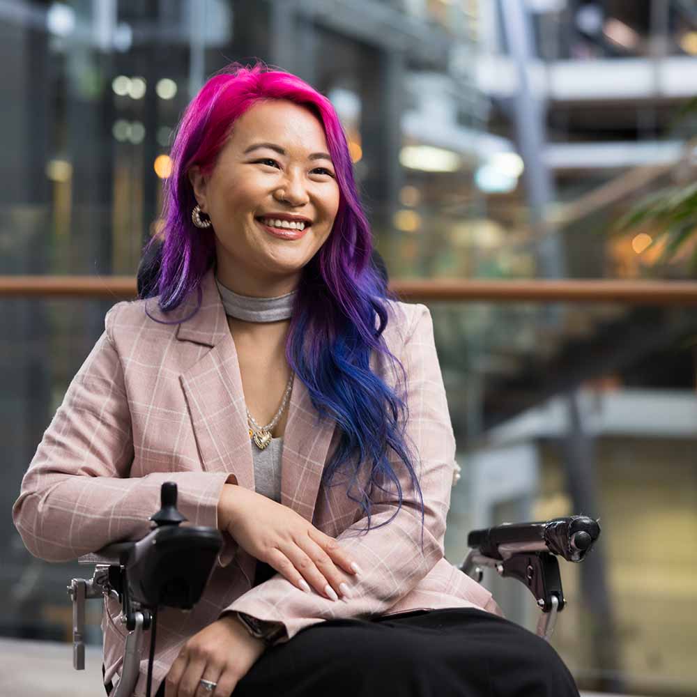 A woman with pink and purple hair sits in a motorised wheelchair.