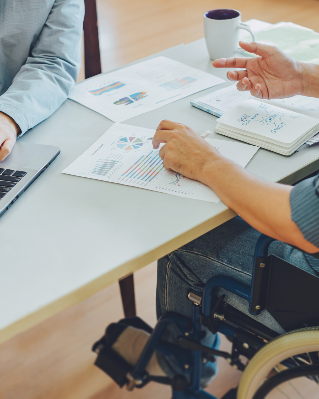 Image of two men sitting at a desk one in a chair and one in a wheelchair. The men are discussing paperwork on the desk in front of them